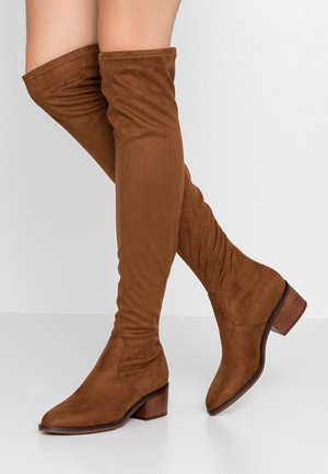 Bottes en daim marron au-dessus du genou avec des talons en bois empilés bas portées sur des jambes croisées devant un fond clair uni.