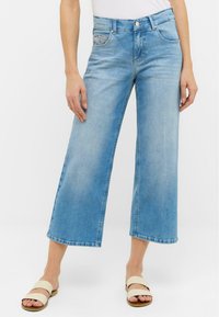 Jeans en denim bleu clair à jambes larges avec une taille haute, un léger délavage et un design de poche décoratif. Associés à des sandales à glisser beiges.
