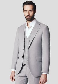 Jasnoszary, dopasowany garnitur z klapą w kształcie notcha, jednorzędowa marynarka i wzorzysta, szara kamizelka pod spodem. Widoczna biała koszula.