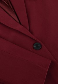 Burgundy blazer featuring a notch lapel, single black button closure, and fine texture. Detail of tailored stitching is visible.