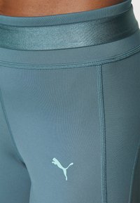 Närbild av turkosa träningsleggings med synliga sömmar och en ljusblå Puma-logotyp på övre låret.