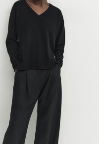 Black knit sweater with a V-neck, relaxed fit, ribbed cuffs, paired with loose-fitting black trousers featuring pleats and side pockets.