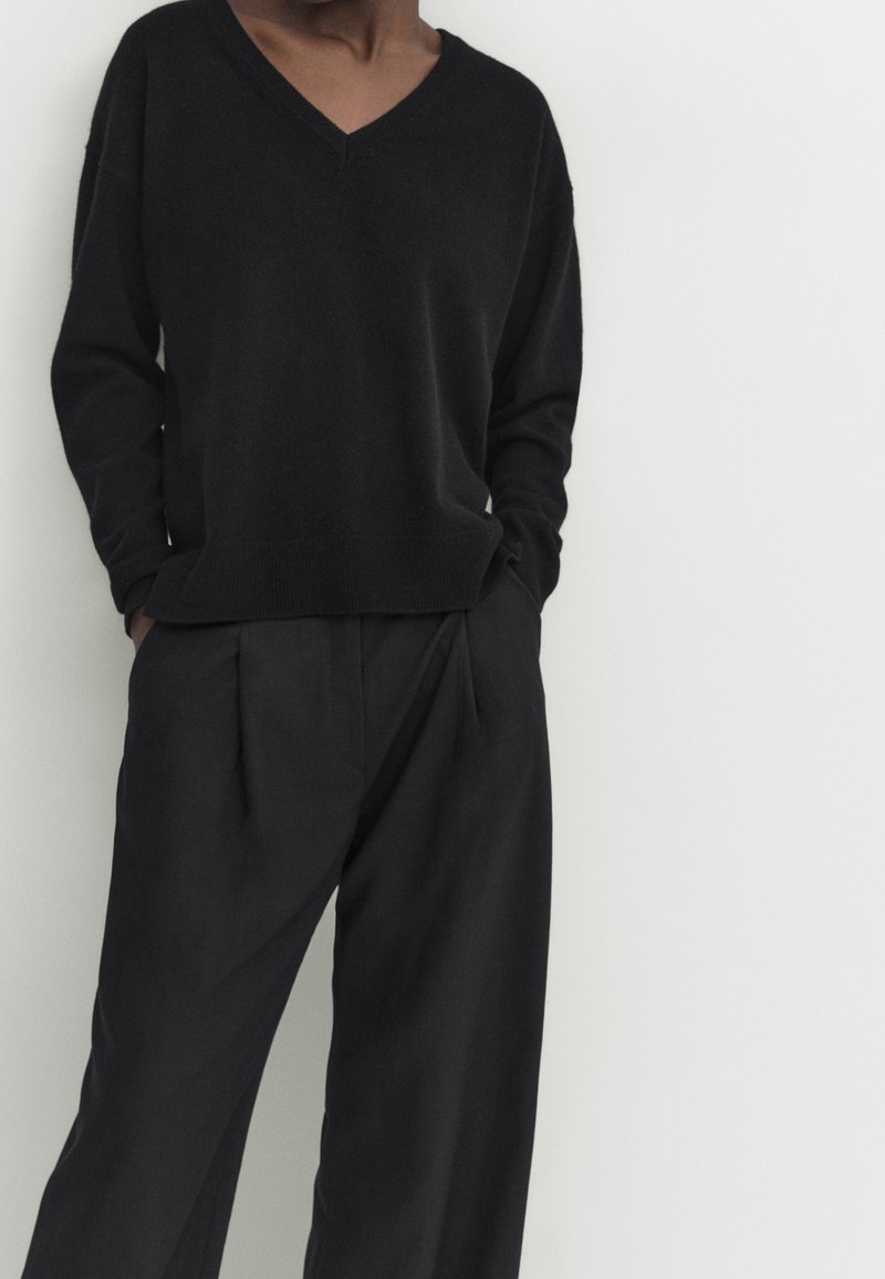 Black knit sweater with a V-neck, relaxed fit, ribbed cuffs, paired with loose-fitting black trousers featuring pleats and side pockets.