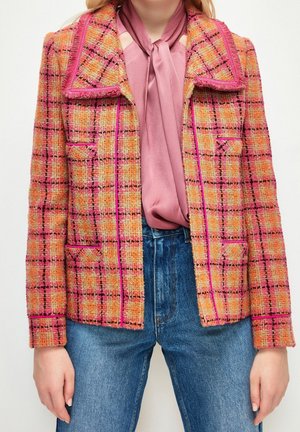 Femme portant une veste à carreaux rose et orange sur un chemisier mauve avec un nœud au col, associée à un jean bleu taille haute.