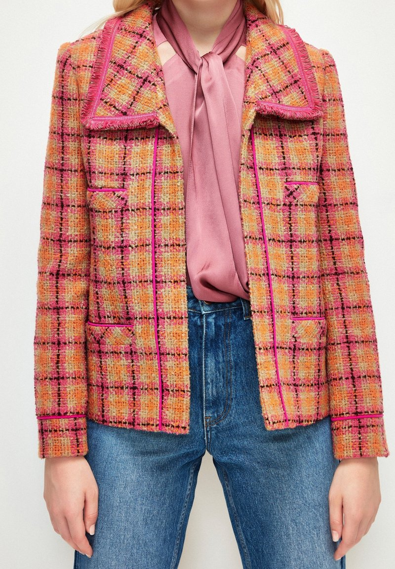 Femme portant une veste à carreaux rose et orange sur un chemisier mauve avec un nœud au col, associée à un jean bleu taille haute.