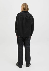 Blonde person wearing black oversized jacket, dark jeans, and black shoes standing facing away on a light gray background.