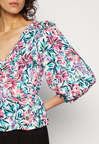 Blusa floral com decote em V, mangas bufantes e padrões vibrantes de rosa, azul e verde num fundo branco. Textura de tecido suave.