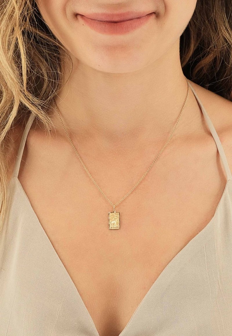 Collier pendentif en or avec un charme rectangulaire présentant un motif embossé, suspendu à une fine chaîne, porté sur un haut de couleur claire.