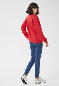 Red sweatshirt with ribbed cuffs and hem, paired with blue denim jeans. Model wears beige sneakers. Background is white, highlighting the outfit.