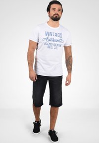 Bela kratka majica s kratkimi rokavi z modrim napisom "VINTAGE Authentic BLEND DENIM REG. CO.", v kombinaciji s temnimi jeans kratkimi hlačami in črnimi supergami.