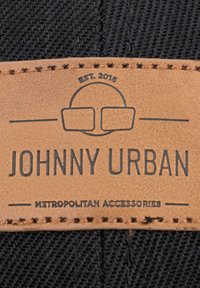 Fekete szövet címke, domború barna bőr folttal. A szöveg tartalmazza a "JOHNNY URBAN," "METROPOLITAN ACCESSORIES" és "EST. 2016" feliratokat, díszöltéssel.