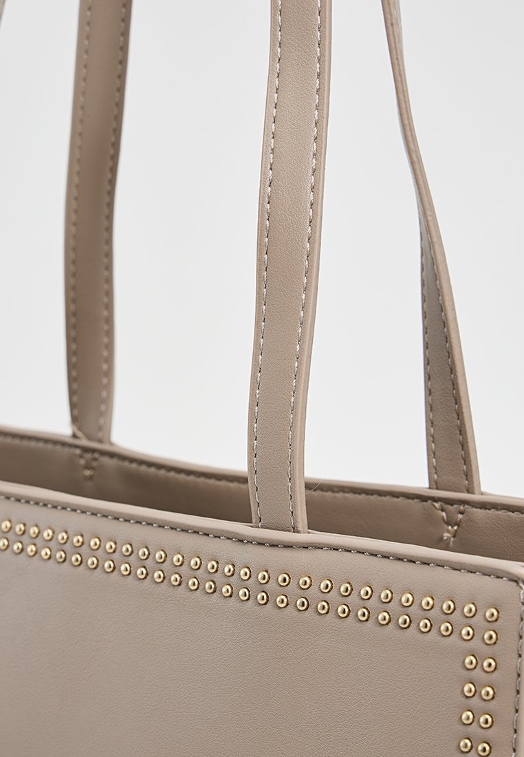 Bolso tote de imitación piel beige con dos asas superiores, que presenta una fila de tachuelas doradas en la parte frontal. Textura suave, diseño estructurado.