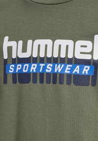 Olijfgroene katoenen T-shirt met grote witte en blauwe tekst "hummel SPORTSWEAR". Het blauwe gedeelte heeft een gestructureerd stippenpatroon.