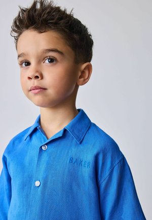 Young boy with short curly hair wearing a blue collared shirt embroidered with "BAKER," looking slightly to the left against a plain background.
