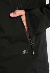 Czarna wodoodporna kurtka z kieszenią na suwak z boku, teksturowanym materiałem oraz małym białym logo przedstawiającym narciarza. Widoczna zapięcie na rzepy przy mankietach.