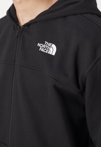 Felpa nera con zip realizzata in tessuto morbido, con logo bianco ricamato di "The North Face" sul lato sinistro del petto.