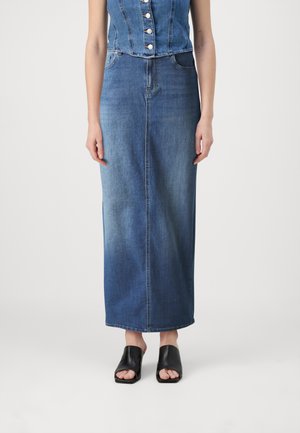 Jupe maxi en denim bleu moyen, coupe droite, avec une couture sur le devant, portée avec des mules ouvertes noires. La texture est lisse.