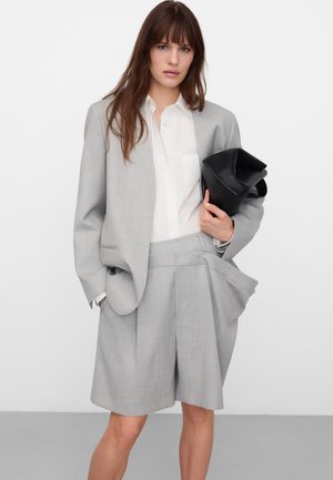 Donna che indossa un blazer oversize grigio chiaro e bermuda plissettati, camicia bianca con bottoni, che tiene una pochette nera su sfondo neutro.