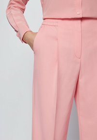 Světle růžová blůza s knoflíkovou přední částí a dlouhými rukávy, spárovaná s odpovídajícími širokými kalhotami s elastickým pasem a bočními kapsami.