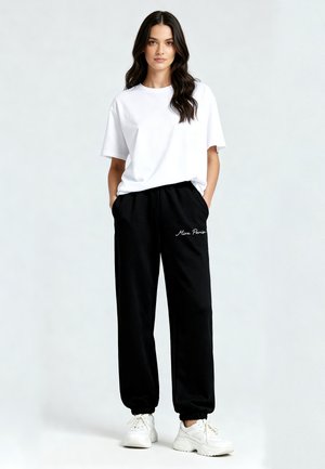 Vrouw draagt een wit T-shirt, zwarte joggingbroek met de tekst "Mino Paris", witte sneakers, staand met handen in de zakken op een effen achtergrond.