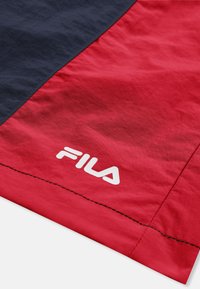Section de tissu rouge et bleu foncé avec le logo blanc FILA imprimé près du bord cousu, probablement sur un matériau de sport ou de vêtements d'activité.
