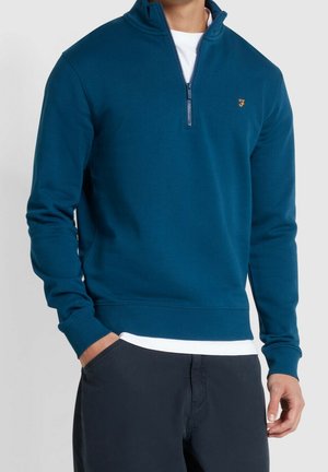 Hombre vistiendo un pulóver de cremallera corta color verde azulado sobre camisa blanca con pantalones azules marinos, con la mano en el bolsillo, posando frente a un fondo liso.