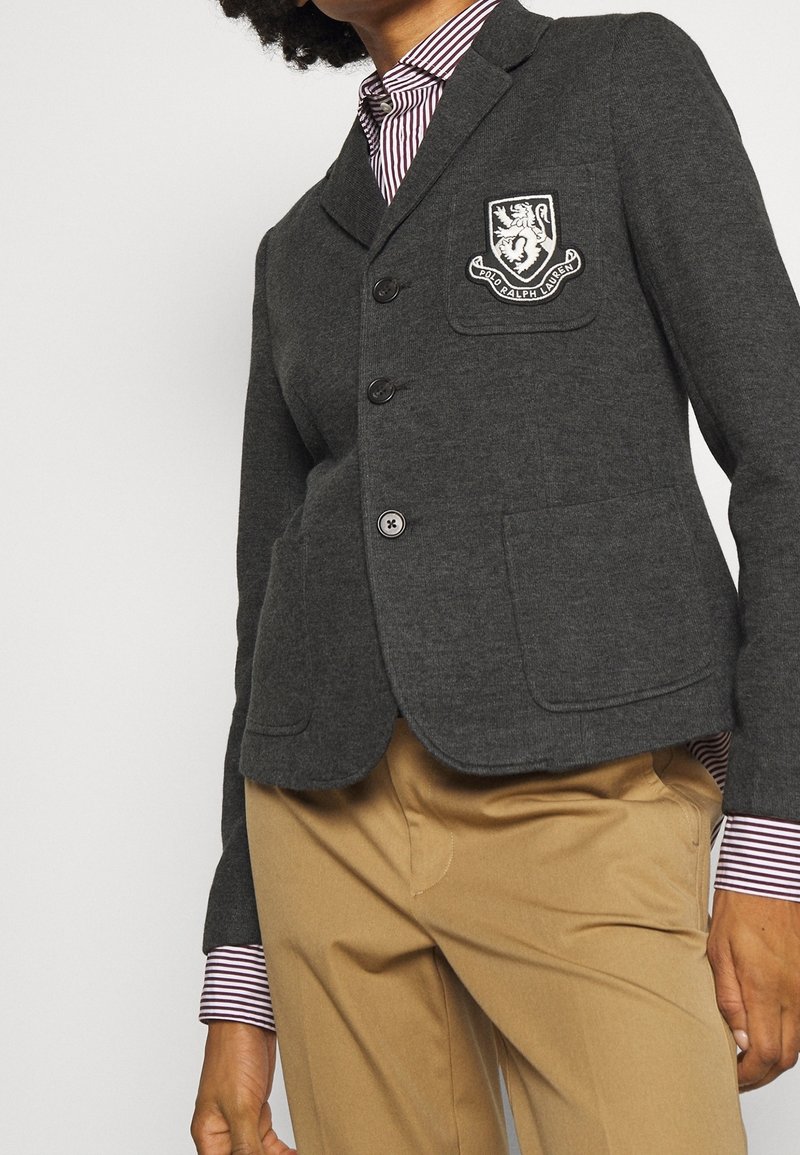 Personne portant un blazer gris avec l'écusson Polo Ralph Lauren, une chemise rayée boutonnée et un pantalon beige, visible de la moitié du torse jusqu'au cou.