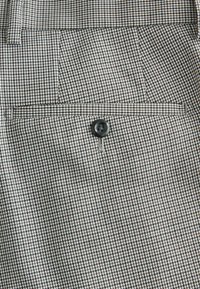 Gros plan sur un pantalon en tissu pied-de-poule marron et blanc avec une poche arrière sécurisée par un seul bouton noir.