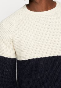 Pull en maille avec moitié supérieure crème et moitié inférieure bleu marine, présentant un motif texturé à tissage ouvert et un col côtelé.
