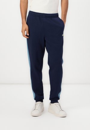 Uomo che indossa pantaloni della tuta blu navy con strisce laterali azzurro chiaro e sneakers bianche, in piedi su uno sfondo bianco semplice.