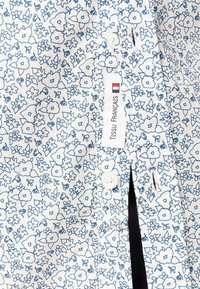 Biała koszula z tkaniny z niebieskim kwiatowym wzorem, z zapięciem na guziki i etykietą z napisem "Tissu Français". Teksturowany, lekki design.