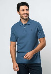 Mies, jolla on tumma tukka ja parta, hymyilee. Hänellä on sininen lyhythihainen poolopaita ja tummat farkut, ja hän seisoo vaaleaa taustaa vasten.