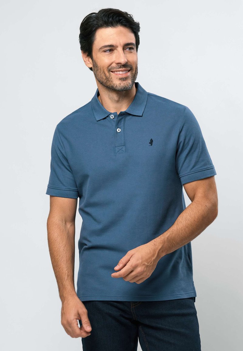 Mies, jolla on tumma tukka ja parta, hymyilee. Hänellä on sininen lyhythihainen poolopaita ja tummat farkut, ja hän seisoo vaaleaa taustaa vasten.