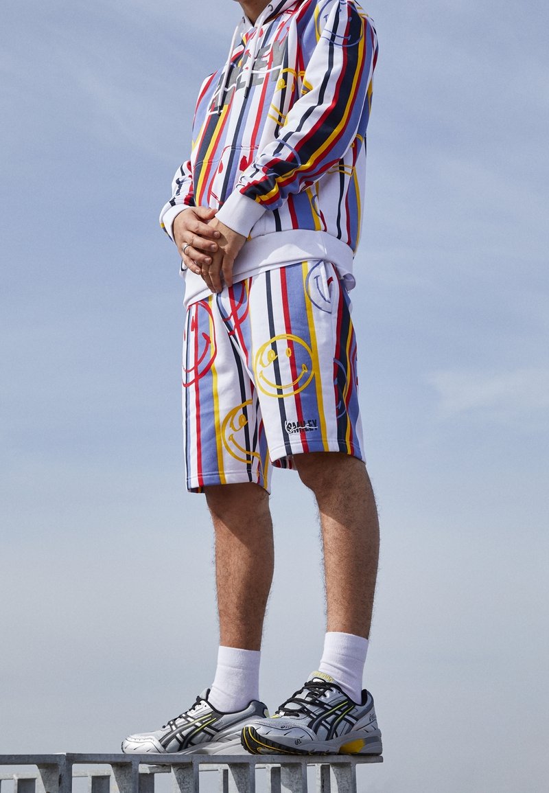 Witte en multikleurige gestreepte hoodie en shorts met glimlachende gezichten. Bevat sportieve schoenen in grijs en zwart met rubberen zolen.