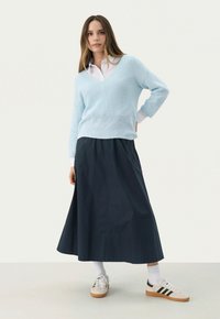 Lichtblauwe, gestructureerde gebreide trui over een witte blouse met kraag, gecombineerd met een donkerblauwe, enkel-lange rok en witte sneakers.
