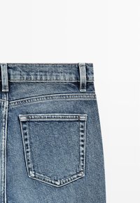 Tył niebieskich dżinsów z widocznym pasem talii, pętlami na pasek oraz pojedynczą prostokątną tylną kieszenią z widocznym przeszyciem.