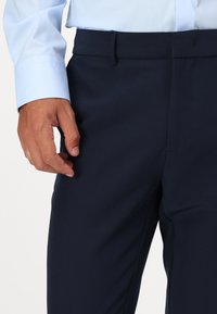 Marineblaue Anzughose mit glatter Textur, gerade geschnitten und mit einem Bund mit dezenter Verschlusstechnik. Die Hand ruht auf dem Stoff.