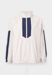 Lagani Nike windbreaker jakna u bijeloj i mornarskoj plavoj boji, koja se ističe visokim ovratnikom, zatvaranjem na patentni zatvarač, elastičnim manžetama i dva bočna džepa.