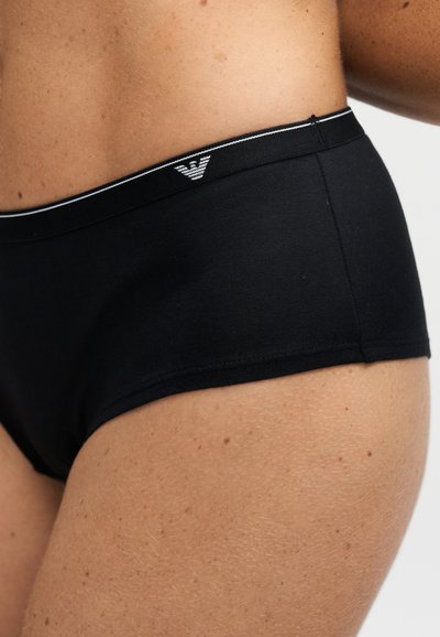 Gros plan d'une personne portant un sous-vêtement noir ajusté avec un petit logo blanc sur la ceinture, sur un fond uni.