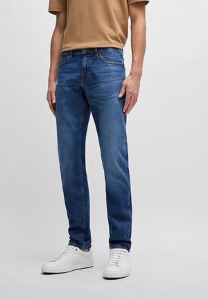 Blue denim jeans with a straight fit, featuring contrasting stitching, five pockets, and a button closure, paired with white sneakers.