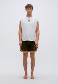 Fehér ujjatlan póló kisméretű fekete grafikával a mellkasán, sötétzöld rövidnadrággal, amely arany színű derékpánttal és logóval van ellátva.