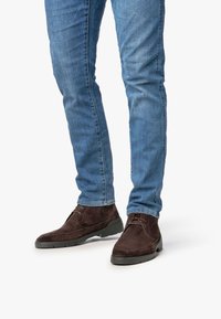 Piernas vistiendo jeans ajustados azules y botas de ante marrón oscuro con cordones hasta el tobillo sobre un fondo blanco liso.