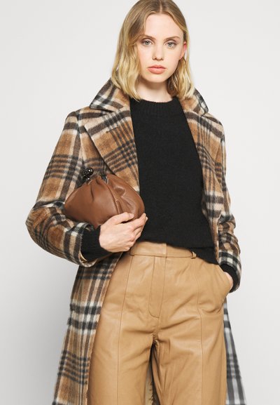 Palton cu ștefănești maro și bej, pulover negru, pantaloni din piele bej, ținând o poșetă din piele maro. Texturi moi cu elemente de design structurat.