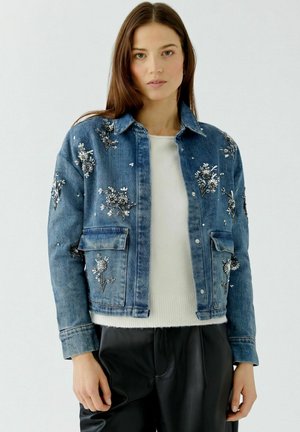 Femme portant une veste en jean bleue avec une broderie florale blanche sur un pull blanc et un pantalon noir, devant un fond clair uni.