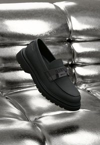 Mocassins noirs en cuir avec une semelle épaisse et chunky, dotés d'une sangle noire brillante avec "REPLAY" embossé. Positionnés sur une surface métallique.