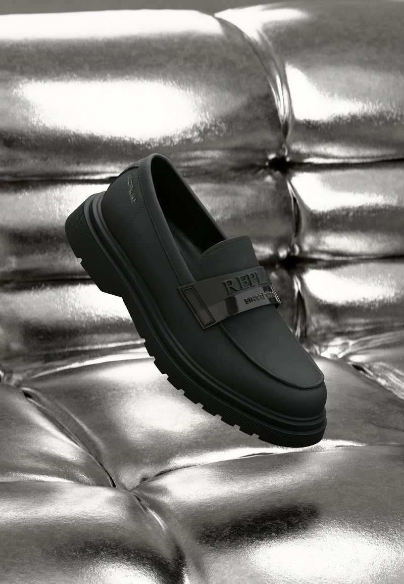 Pantofi loafers negri din piele, cu o talpă groasă și masivă, având o curea lucioasă neagră cu "REPLAY" embosat. Poziționați pe o suprafață metalică.