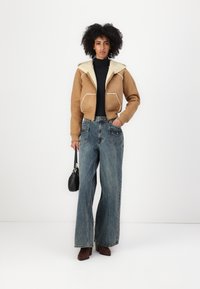 Veste en daim marron cropped avec un col en polaire crème, associée à un jean bleu clair à jambes larges. Sac à main noir et bottines marron.