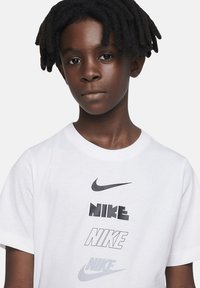Camiseta de algodón blanca con múltiples logotipos de Nike en diferentes tonos de gris. Manga corta y diseño de cuello redondo. Textura suave.