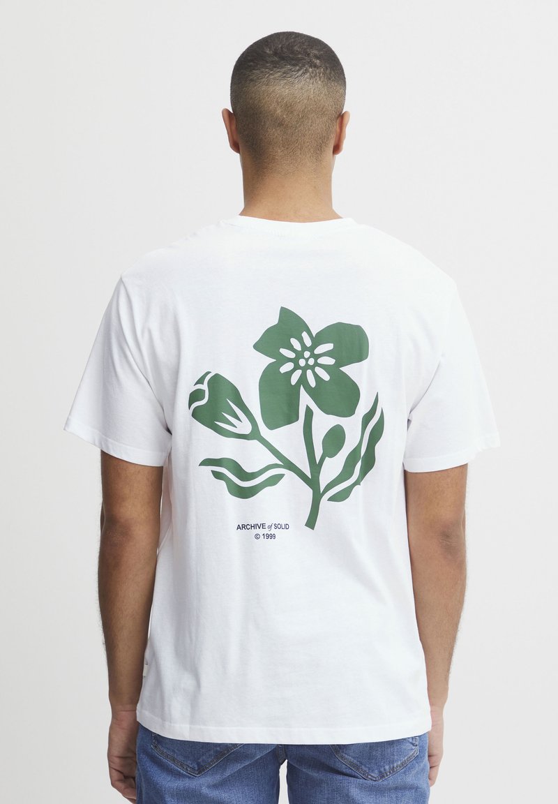 Camiseta de algodón blanca con un estampado floral verde en la parte trasera, que incluye hojas y pétalos estilizados. El texto en la parte inferior dice "ARCHIVO / VENDIDO © 1999."