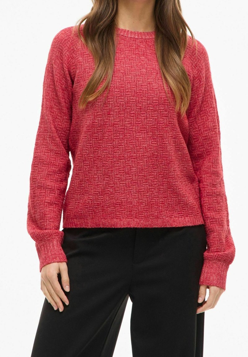 Femme portant un pull rouge texturé en maille à manches longues et un pantalon noir, debout avec les mains reposant le long du corps.